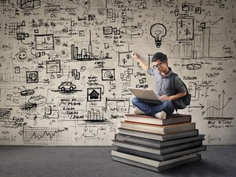 Image depicting a student sitting on top of a pile of books while working on a computer