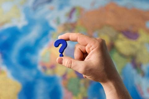 A hand holding up a question mark in front of a world map