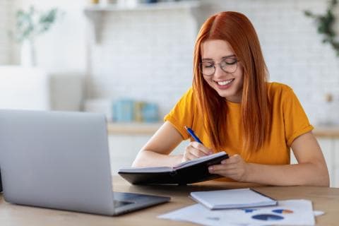 Student at a laptop smiling with a notebook in her hand