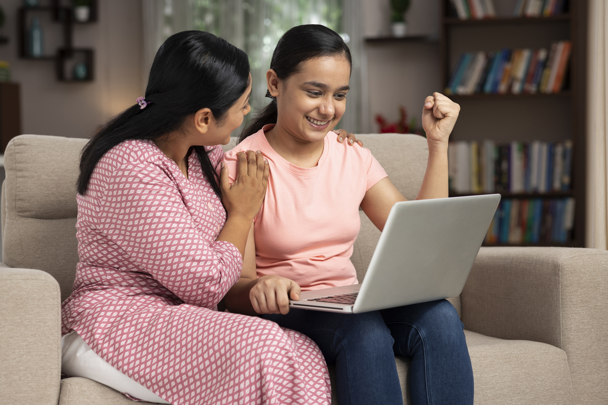 South Asian school girl cheering at computer screen, with supportive mother next to her