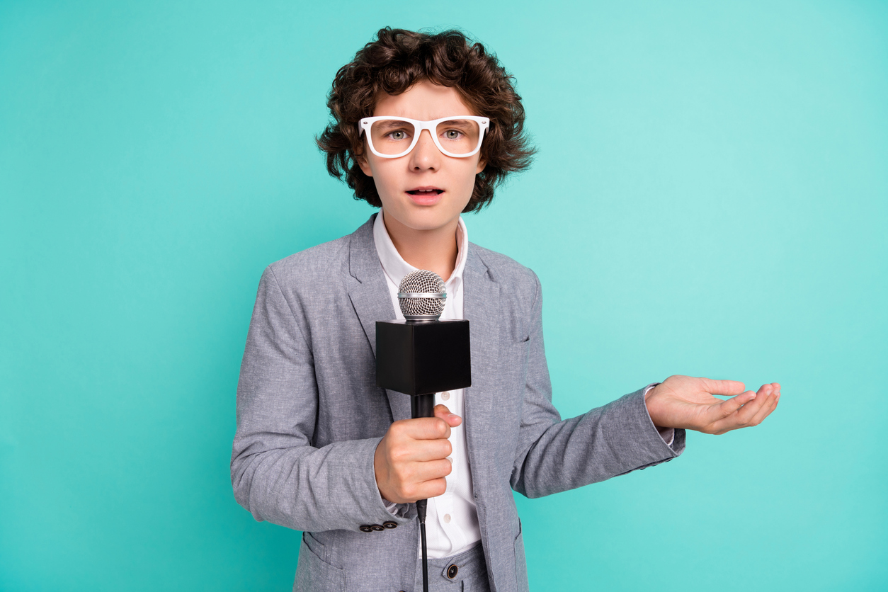 Student dressed in suit, wig and outsized glasses, holding a microphone and pretending to report to camera