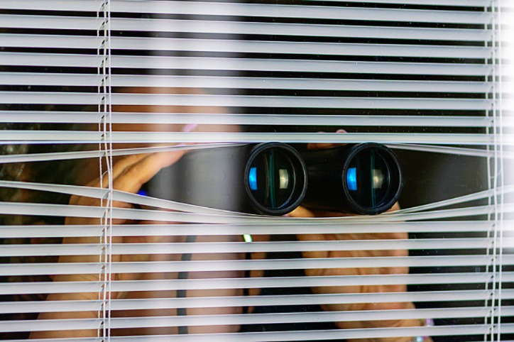 A woman peeking through blinds while holding binoculars, illustrating suspicion