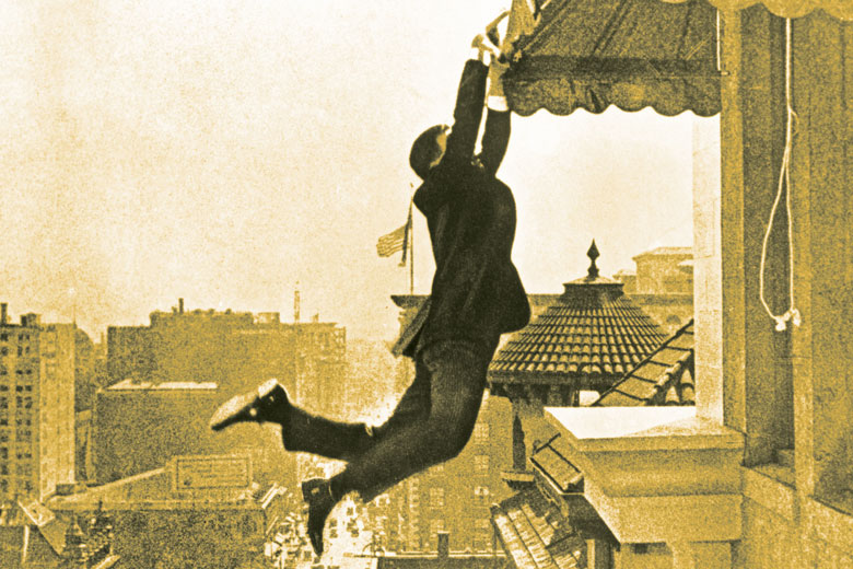 A man hanging onto a window shade outside a building high above a street.