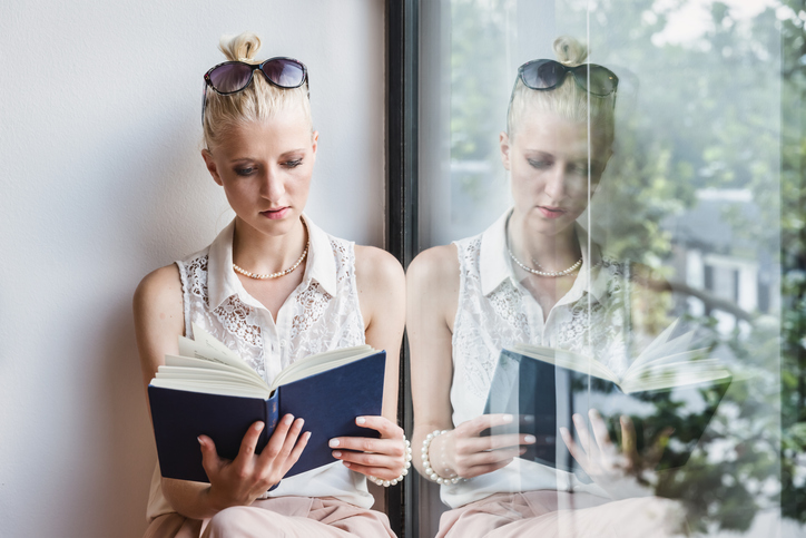 A woman reads a book on a window bench, with a reflection of her doing so in the window, illustrating duplicate publication reading a book