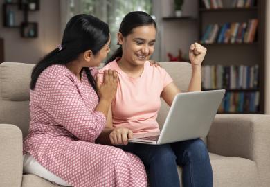 South Asian school girl cheering at computer screen, with supportive mother next to her