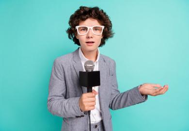 Student dressed in suit, wig and outsized glasses, holding a microphone and pretending to report to camera