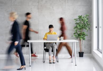 Woman sits at desk, while people rush all around her