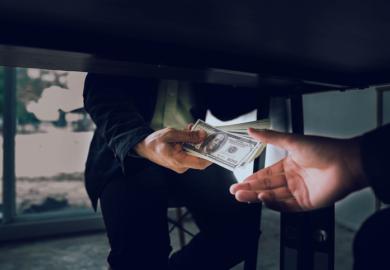 A man passes hundred-dollar bills under the table, illustrating corruption