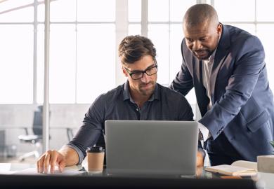 A man points to a computer screen to explain something to another man 