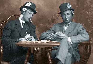 Two young men pretending to play cards. They are smoking and they sit close to a small table. Both men wear hats and have their legs crossed. One man is peeking into the other's card hand. Both guys have aces, kings and other cards visibly hidden in all p