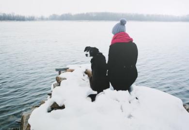 Woman and dog sitting on a snowy river bank 