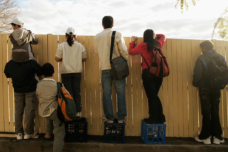 Members of the public stand on crates to look over a fence in Sydney, Australia. To illustrate structures put in place by universities to limit the salary and progression of specialists compared with academics.