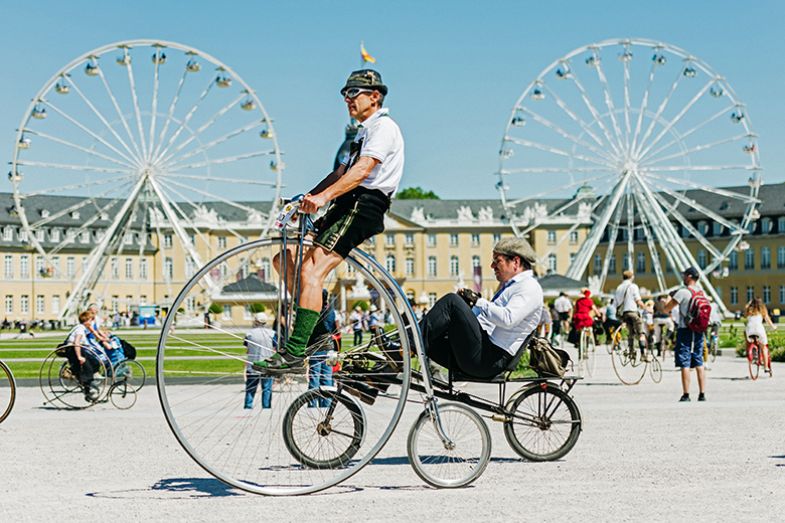 Participants dressed in historical clothing ride high-wheel bicycles during a bicycle event at Schloss Karlsruhe, Germany, with ferris wheels in the background. To illustrate competition of universities to succeed in excellence initiatives. Participants dressed in historical clothing ride high-wheel bicycles during a bicycle event at Schloss Karlsruhe, Germany, with ferris wheels in the background. To illustrate competition of universities to succeed in excellence initiatives.