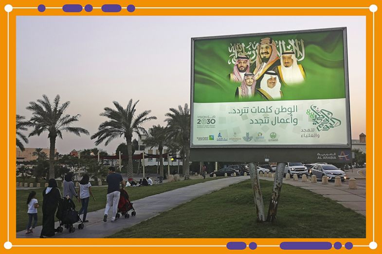 Pedestrians walk past an illuminated sign showing portraits of the Saudi Royal family and Vision 2030 branding on a street in Dhahran, Saudi Arabia, 