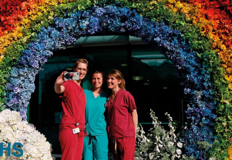 Floral rainbow archway tribute to NHS staff during the COVID-19 pandemic with three Doctors taking a selfie photograph