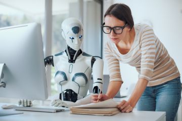 AI robot and woman marking paper AI robot sat at a computer, while a woman in glasses sits next to pointing at a paper