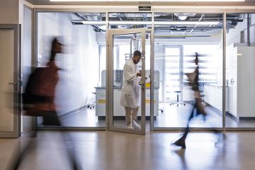 A chemical worker in a white coat stands between the doors of the faculty laboratory, students rush past him in the corridor