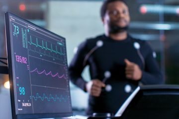 Close-up Shot of a Monitor With EKG Data. Male Athlete Runs on a Treadmill with Electrodes Attached to His Body while Sport Scientist Holds Tablet and Supervises EKG Status in the Background Close-up Shot of a Monitor With EKG Data. Male Athlete Runs on a Treadmill with Electrodes Attached to His Body while Sport Scientist Holds Tablet and Supervises EKG Status in the Background