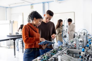 A male and female student using a digital tablet while operating a production line robot in a lab.