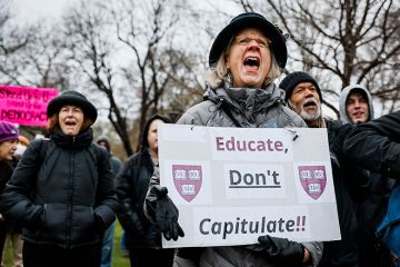 A protester holds a sign reading "Educate, Don't Capitulate!!" during a rally at Cambridge Common to urge Harvard to resist President Trump's influence on the institution, 12 April 2025. A protester holds a sign reading "Educate, Don't Capitulate!!" during a rally at Cambridge Common to urge Harvard to resist President Trump's influence on the institution, 12 April 2025.