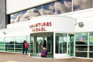 Person pulling a suitcase entering terminal building at Cardiff Wales Airport Cardiff, Wales - June 2019:: Person pulling a suitcase entering the terminal building at Cardiff Wales Airport. Blue sky and clouds are reflected in the windows. The airport is publicly owned by the Welsh Government.
