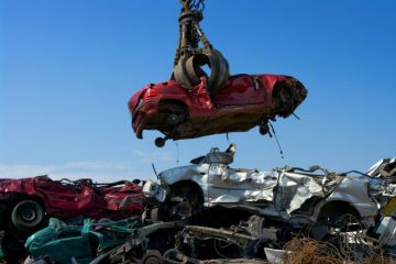 A shattered car is lowered into a pile of other crushed cars