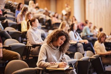 Female student writing in notebook in busy lecture theatre