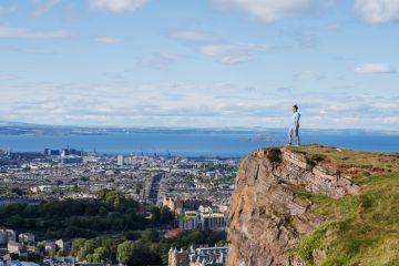 Standing on a rocky precipice, a person overlooks urban scenery and the ocean, under a bright sky Standing on a rocky precipice, a person overlooks urban scenery and the ocean, under a bright sky