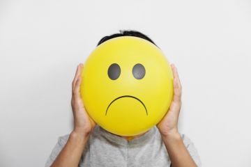 Someone holds a frowning emoji face in front of their own Someone holds a frowning emoji face in front of their own