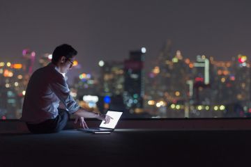 A man in a dark room overlooking a cityscape at night uses a laptop A man in a dark room overlooking a cityscape at night uses a laptop