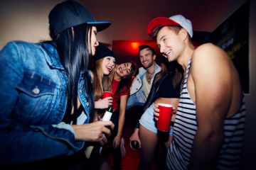 Low angle group of young stylish people standing in dark hall of nightclub together, talking, laughing and drinking beer as they wait to be admitted
