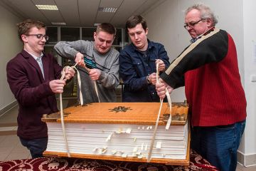 Men struggling to lift a giant book. To illustrate that three major research universities have opted out of new Elsevier deal amid concerns over the cost.