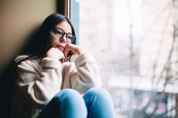 A woman sits by a window looking sad