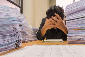 An academic with their head on their desk surrounded by exam scripts
