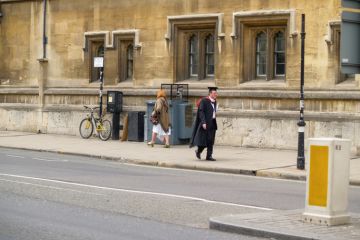 Oxford United Kingdom - June 19 2009; University city street scene with man in gown and mortarboard or Oxford cap walking on sidewalk. Oxford United Kingdom - June 19 2009; University city street scene with man in gown and mortarboard or Oxford cap walking on sidewalk.