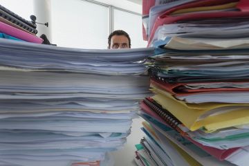 A man peers out from behind a pile of paperwork
