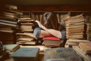 A young researcher with her head on her desk surrounded by piles of documents, illustrating early-career overwork A young researcher with her head on her desk surrounded by piles of documents, illustrating early-career overwork