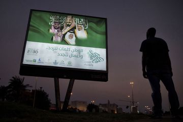 A pedestrian pauses near an illuminated sign showing portraits of the Saudi Arabian Royal family advertising Vision 2030 in Dhahran, Saudi Arabia. A pedestrian pauses near an illuminated sign showing portraits of the Saudi Arabian Royal family advertising Vision 2030 in Dhahran, Saudi Arabia.