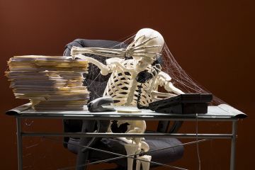Skeleton sitting at desk on telephone, with webs and stacks of paperwork. To illustrate that colleges are in limbo amid ‘two-year wait’ for degree-awarding powers.