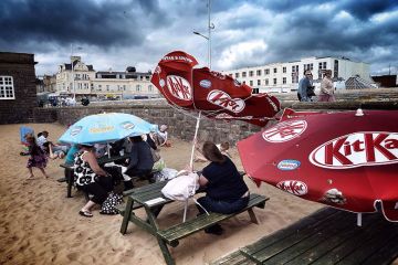 Holidaymakers brave the weather and shelter under an umbrella as another turns inside out in the wind, on the beach in Weston-super-Mare, England. To illustrate a summer of ‘chaos’ being predicted as mass job cuts close the academic year. Holidaymakers brave the weather and shelter under an umbrella as another turns inside out in the wind, on the beach in Weston-super-Mare, England. To illustrate a summer of ‘chaos’ being predicted as mass job cuts close the academic year.