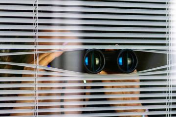A woman peeking through blinds while holding binoculars, illustrating suspicion A woman peeking through blinds while holding binoculars, illustrating suspicion