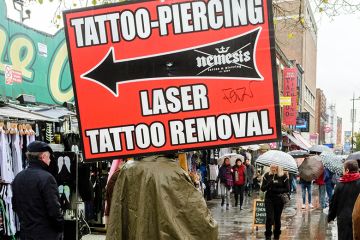 Person holding sign advertising tattoo, piercing and tattoo removal services, Camden High street, London, UK