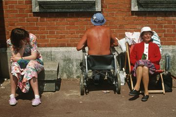 Ageing, elderly parents sunbathe with a teenage daughter as the father oddly faces a brick wall while sat in his wheelchair. Looking bored with the family holiday, the young lady of about 18 years of age, sits on a concrete block