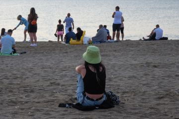 The young woman sits alone on the beach and admires the sea. In front of her are several people on the beach. Romania, Costinesti. August, 19, 2022