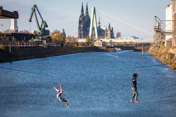man and woman on slacklines in the Rhine harbour in Deutz, with the cathedral and Severins bridge in the background, Cologne, Germany, as a metaphor for European universities balancing cost and ambition