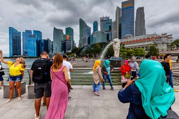 Tourists of various nationalities pose for photos in front of the Merlion statue and Singapore skyline, illustrating Singapore’s international nature