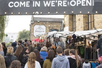 crowds passing under an entrance sign reading " Come in We're (very) Open.