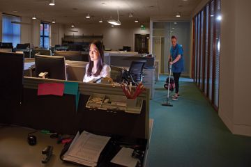 A young woman has stayed behind to get her work done in a large open plan office . A cleaner is vacuuming behind her .to illustrate ‘Illegal’ terms and conditions add to academic overwork crisis