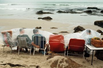 People on deckchairs shelter under plastic sheets on beach