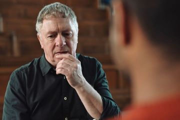Man with grey hair and black shirt holding hand at mouth whilst thinking and talking to male Man with grey hair and black shirt holding hand at mouth whilst thinking and talking to male